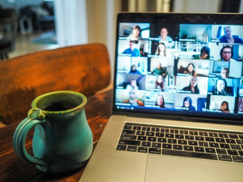 Open laptop screen displays many people on virtual video call; laptop is on table near a mug of coffee