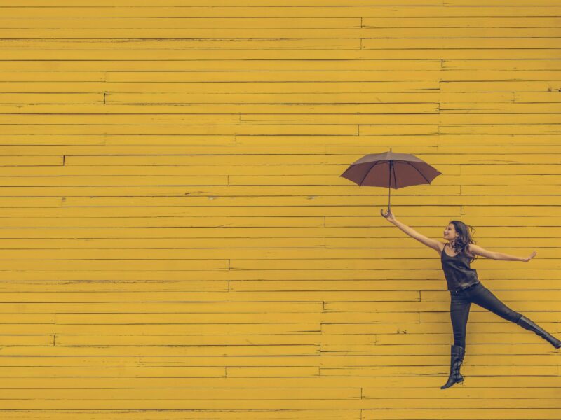 Woman appears to be floating holding an umbrella against a yellow brick wall