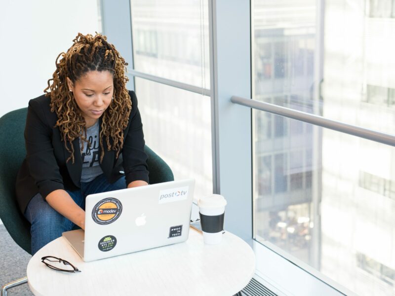 Woman in black blazer works on laptop next to tall window