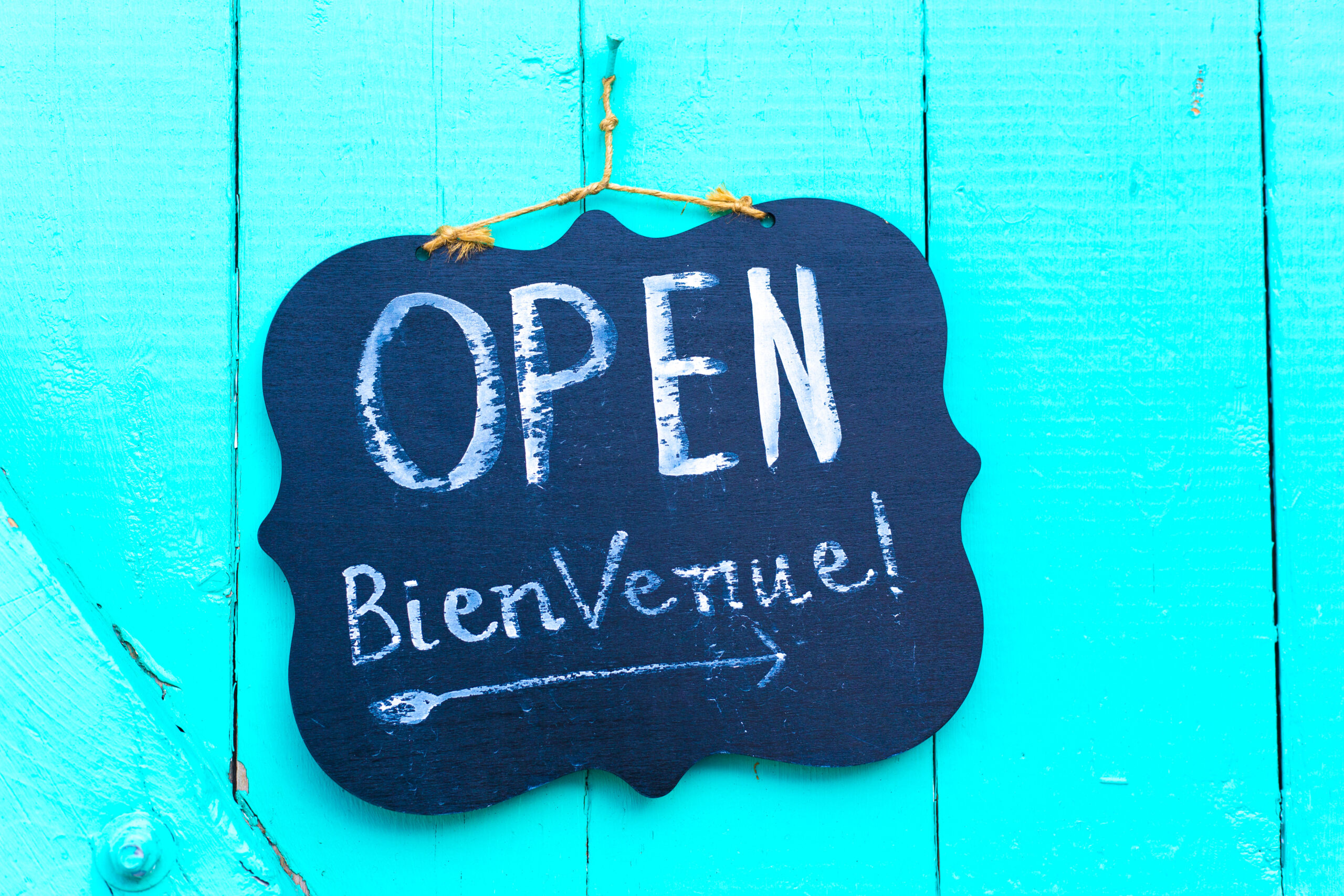 Blue sign says Open and Bienvenue, hanging against a distressed turquoise wood wall